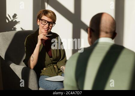 Young woman in eyeglasses sitting on armchair and talking to the man they have an interview at office Stock Photo