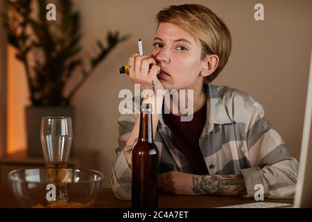 Portrait of young woman with short hair sitting at the table with beer and holding cigarette and looking at camera Stock Photo