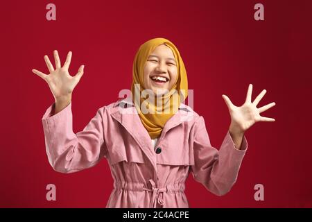 Portrait of happy Asian muslim teenage girl smiling and waving at camera, wearing red hijab against red background Stock Photo