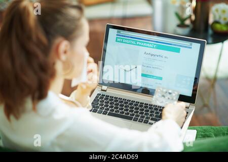 Seen from behind ill woman in white blouse with empty blister pack of pills ordering medicine online on a laptop in the house in sunny day. Stock Photo