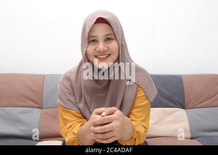 Beautiful Asian muslim lady doing selfie portrait on phone or doing video call while sitting on sofa, happy smiling cheerful expression Stock Photo
