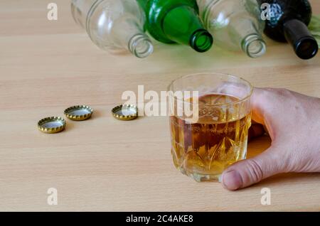 The social problem of home alcoholism concept. Male hand holds a glass with beer on the background of empty drunk bottles of various alcohol.An adult Stock Photo