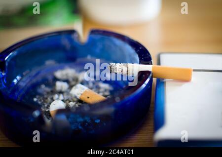 burning cigarette  in a blue glass ashtray with cigarette butters and ash pile isolated on table, selective focus Stock Photo