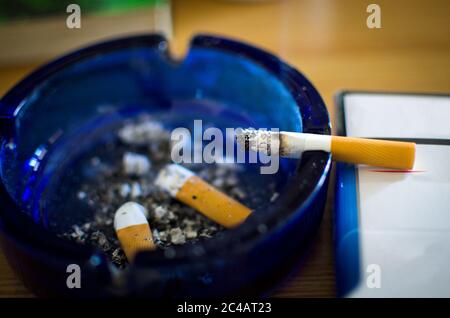 burning cigarette  in a blue glass ashtray with cigarette butters and ash pile isolated on table, selective focus Stock Photo