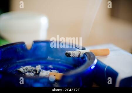 burning cigarette  in a blue glass ashtray with cigarette butters and ash pile isolated on table, selective focus Stock Photo
