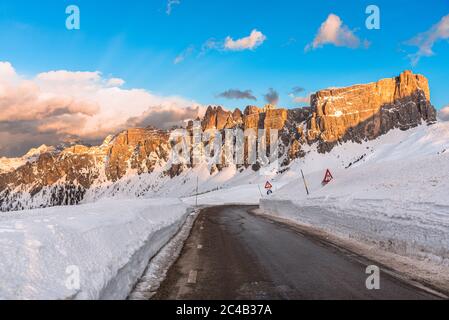 Empty straight stretch of mountain pass road in a magnificent snowy landscape with towering rocky peaks warmly lit by a setting sun in winter Stock Photo