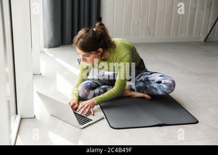 Photo of focused brunette sportswoman using laptop while working out on mat in cozy room Stock Photo