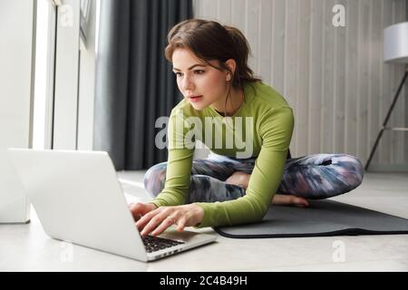Photo of focused brunette sportswoman using laptop while working out on mat in cozy room Stock Photo