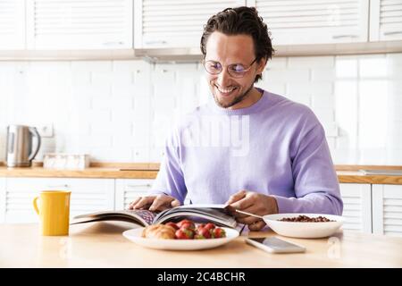 Attractive happy young man sitting at the kitchen table, having breakfast, eating cereal, reading magazine Stock Photo