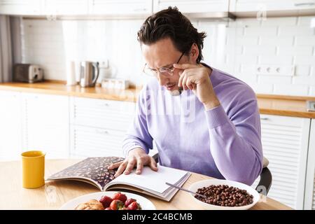 Attractive concentrated young man sitting at the kitchen table, having breakfast, eating cereal, reading magazine Stock Photo