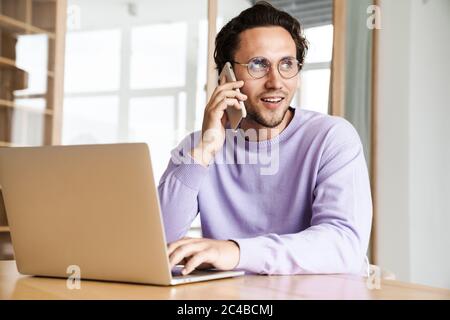 Attractive cheerful young man sitting at the kitchen table, working on laptop computer, talking on mobile phone Stock Photo