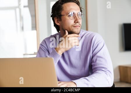 Attractive pensive young man sitting at the kitchen table, using laptop Stock Photo