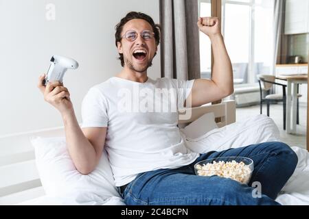 Handsome cheerful young man playing on playstation while laying on bed at home, eating popcorn, celebrating Stock Photo