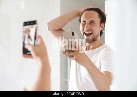 Attractive happy confident young man standing in front of the bathroom mirror, taking a selfie Stock Photo