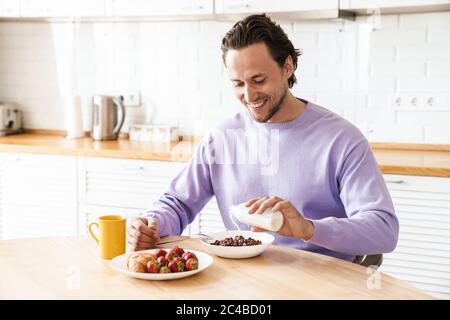 Attractive happy young man sitting at the kitchen table, having breakfast, eating cereal Stock Photo