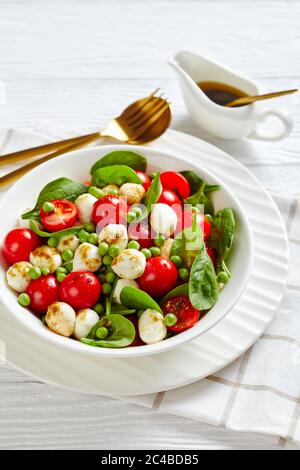 Simple veggie salad of young peas with grape tomatoes, fresh baby spinach leaves, mini mozzarella balls, balsamic vinegar sauce on a white wooden back Stock Photo