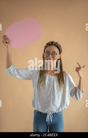 Happy young African American female in eyeglasses holding pink dialogue cloud, pointing her index finger at it Stock Photo