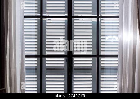 gray wooden window shutters protect the room from excessive sunlight, a window with closed shutters and open curtains of white textiles. Stock Photo