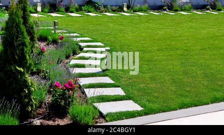 backyard garden with a flower bed with roses and lavender with a path of marble tiles on the meadow with a green lawn on a sunny day. Stock Photo