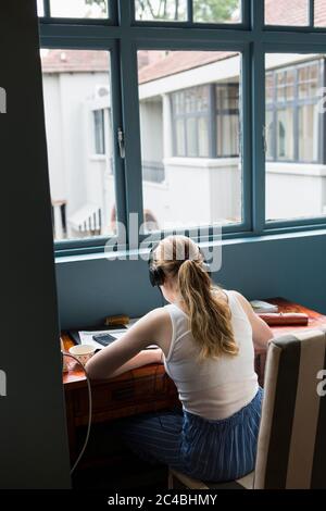 A thirteen year old girl wearing headphones doing homework seated in a quiet spot. Stock Photo