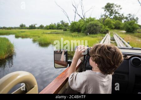 A six year old boy using binoculars looking over water from a safari vehicle Stock Photo