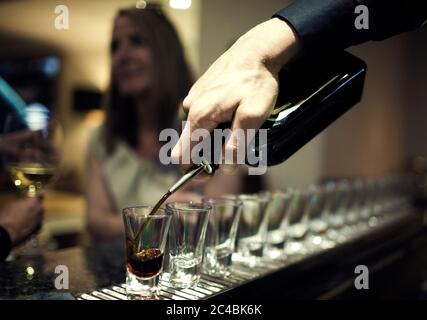 Close up of bartender pouring drinks from bottle into a row of shot glasses standing on bar counter, woman sitting in background. Stock Photo