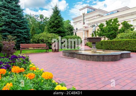 a stone fountain with a sculpture in the backyard with a wooden bench and an urn surrounded by a garden of green plants and a flower bed with flowers. Stock Photo