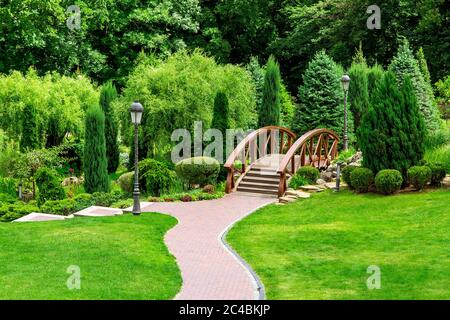 Landscaping of a park in the backyard with a path for walks through the garden through a wooden decorative bridge to bushes and trees. Stock Photo