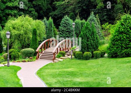 Landscaping of a park in the backyard with a footpath for walks through the wooden decorative bridge to bushes and trees on summer day. Stock Photo