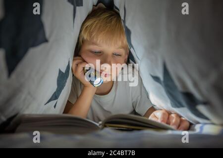 A little boy reads a book with a flashlight under the covers at night. Stock Photo
