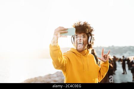 Young Afro man taking self video while listening music with wireless headphones - Happy mixed race guy having fun with new technology apps Stock Photo