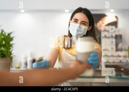 Bar owner working only with take away orders during corona virus outbreak - Young woman worker wearing face surgical mask giving coffee to customer Stock Photo