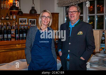 Journalist Angela Berg stands smiling next to a man with glasses in a dark grey traditional Trachten jacket in front of a bar counter with wine bottles in the Hotel and Restaurant Kronenschlösschen in Hattenheim, Eltville am Rhein, Hesse, Germany. The man has his hands in his trouser pockets and wears a patterned pocket square. Stacked plates stand on a table with a white cloth on the right. On the evening of 25 February 2017 two dinners of the 21st Rheingau Gourmet & Wein Festival take place in the house: the gala dinner of star chef Rodolfo Guzmán with host chef Simon Stirnal and the "Vega S Stock Photo