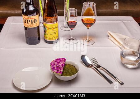 The dessert "Die Rose des Jahres" ("the rose of the year") by star chef Rodolfo Guzmán (Restaurant Boragó, Santiago de Chile) stands in a white bowl on a laid table in the Hotel and Restaurant Kronenschlösschen in Hattenheim, Eltville am Rhein, Hesse, Germany. A semicircular white piece topped with violet petals (possibly rose petals) leans against the rim of the bowl above a green, powdery layer. Behind it stand a bottle of Ramos Pinto RP10, a ten-year-old tawny port from Quinta de Ervamoira, and a bottle of 1989 Hattenheimer Nussbrunnen Riesling Auslese from the Schloss Schönborn estate, wit Stock Photo