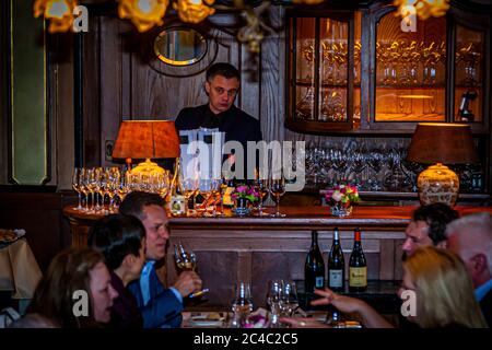 Guests of the lobster lunch sit with white wine glasses at tables in front of the bar of the Hotel and Restaurant Kronenschlösschen in Hattenheim, Eltville am Rhein, Hesse, Germany. A young man in a dark suit stands behind the wood-panelled bar holding white cards. Rows of wine glasses, flower arrangements and two lamps with orange shades stand on the counter. Three bottles of the wine pairing, including an Alvarinho Soalheiro, stand on a sideboard on the right. Journalist Angela Berg sits in the front right. The wine tasting is part of the lobster lunch of the 21st Rheingau Gourmet & Wein Fes Stock Photo