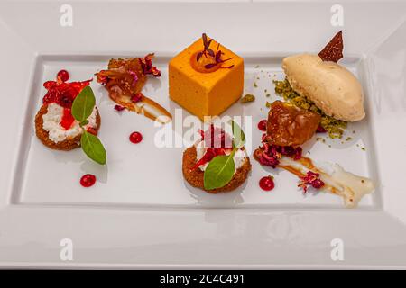 An orange cube topped with purple cress stands in the centre of a rectangular white plate. Around it lie two biscuit rounds with white cream, red fruit strips and basil leaves, glazed fruit pieces with caramel sauce and dried petals, and dots of red jelly. On the right a quenelle of ice cream flecked with vanilla rests on green crumbs, with a crisp. The gourmet dish (possibly a dessert) belongs to the 21st Rheingau Gourmet & Wein Festival at the Hotel and Restaurant Kronenschlösschen in Hattenheim, Eltville am Rhein, Hesse, Germany. On 10 March 2017 two festival events take place in the house  Stock Photo