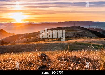 Landscape in Santa Cruz mountains, with sun rays illuminating hills covered in dry grass; People visible on the hiking trail, watching the sunset; San Stock Photo