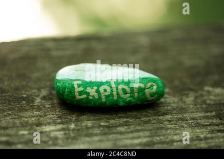 A kindness rock that reads Explore in gold paint with a blue and green design sits on a wooden fence in the arboretum in Acton, Massachusetts, USA. Stock Photo