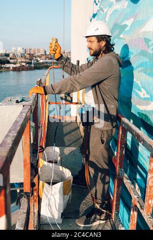 Contractor, builder at high altitude in a construction cradle presses a button to knock from a height to the ground against the background of the city Stock Photo