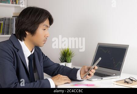 Young Asian Forex Trader or Investor or Businessman in Suit Looking Smartphone and Trading Forex or Stock Chart by Laptop in Trader Room Stock Photo