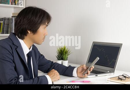 Young Asian Forex Trader or Investor or Businessman in Suit Hold Smartphone and Trading Forex or Stock Chart by Laptop in Trader Room Stock Photo