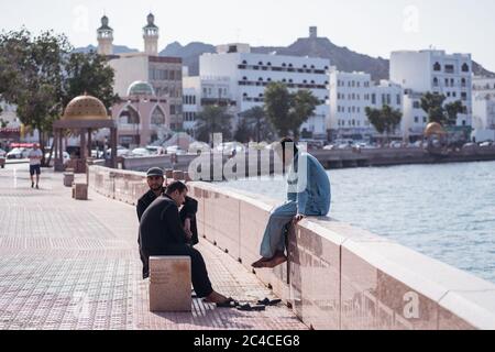 Muscat / Oman - February 15, 2020: group of Muslim male friends enjoy leisure time sitting in the Corniche of Muscat with Mutrah Souq in the background Stock Photo