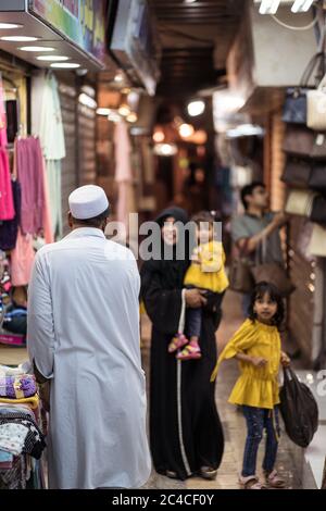Muscat / Oman - February 15, 2020: Muslim woman wearing black abaya shopping in Mutrah Souq with two young daughters and shop seller in white traditional clothing Stock Photo
