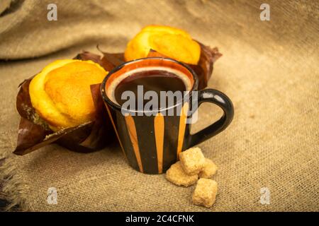 Two fresh cupcakes, a mug of coffee, and chunks of brown cane sugar on a rough-textured homespun cloth. Close up Stock Photo