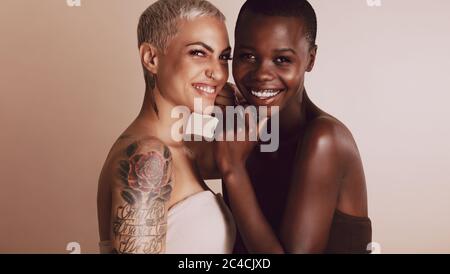 Two beautiful women standing together against beige background. Female models of different ethnicities with short hairstyle looking at camera and smil Stock Photo
