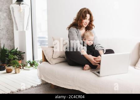 Young beautiful woman sitting on sofa with her little handsome son dreamily using laptop together. Mom with baby boy happily spending time in living Stock Photo