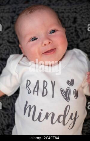 A newborn happy baby girl laying on a dark rug with a white baby vest with the words Baby Mindy. Photo by Sam Mellish Stock Photo
