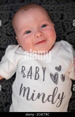 A newborn happy baby girl laying on a dark rug with a white baby vest with the words Baby Mindy. Photo by Sam Mellish Stock Photo