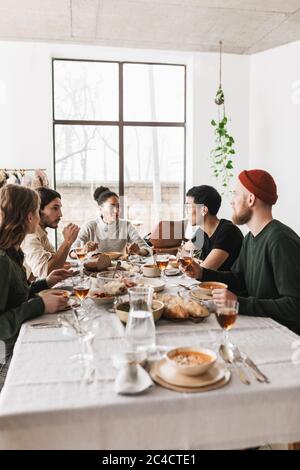 Group of attractive international friends sitting at the table full of food dreamily talking together. Young colleagues having lunch happily spending Stock Photo