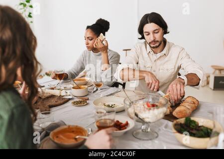 Thoughtful man with dark hair and beard sitting at the table cutting bread while afro american woman near dreamily eating. Group of young Stock Photo